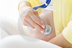 One european pregnant woman with smile is listening heart beat of unborn child inside her tummy by stethoscope while she