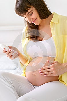 One european pregnant woman with smile is using moisturizing cream on her tummy for prevent stretch marks from pregnancy while she