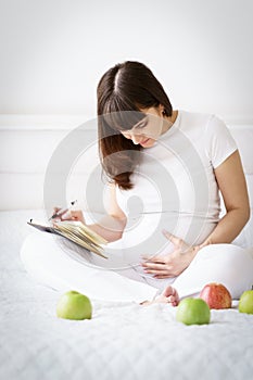 One european pregnant woman writes some idea or thouths in note book by pen sitting on white bed of light room at sunny day.