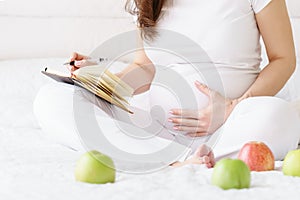 One european pregnant woman writes some idea or thouths in note book by pen sitting on white bed of light room at sunny day.