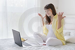 One european pregnant woman with yelow shirt has much emotions from screen of laptop while she is sitting on bed in white bedroom