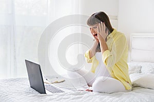 One european pregnant woman with yelow shirt has much emotions from screen of laptop while she is sitting on bed in white bedroom