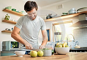 One fit young caucasian man cutting ingredients with knife to make healthy green detox smoothie while wearing earphones