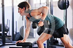 One fit young caucasian man doing single arm bent over rows on a bench with dumbbells while training in a gym. Focused