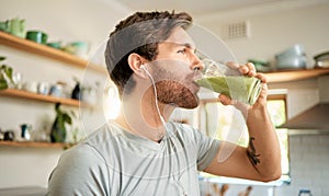 One fit young caucasian man drinking a glass of healthy green detox smoothie while wearing earphones in a kitchen at