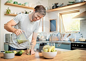 One fit young caucasian man pouring healthy green detox smoothie from blender while wearing earphones in kitchen at home