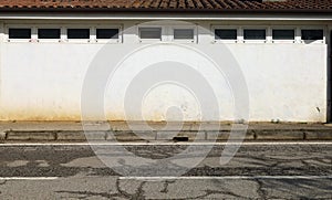 One floor building with white wall and row of small windows. Earthenware tiles rooftop. Concrete sidewalk and road in front