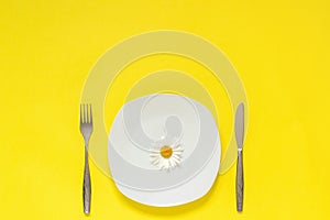 One flower chamomile daisy on plate, cutlery fork knife on yellow paper background Concept vegetarian healthy eating diet or