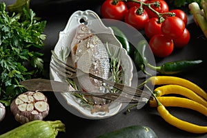 Fresh raw dorada fish in a white dish with a set of vegetables on a black table