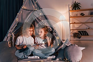 One girl holding flashlight, other reading the book. Two little friends is in the tent in domestic room together