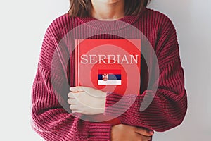 one girl schoolgirl in burgundy sweater holds red serbian language textbook with Serbia flag. foreign language learning concept photo