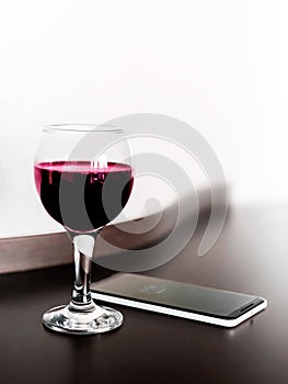 One glass of red wine and a mobile phone on brown wodden table and white blurred wall background. Alcohol drink and smartphone on