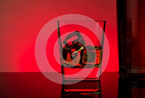 One glass of whiskey with ice cubes near bottle on table with reflection, red lights disco atmosphere