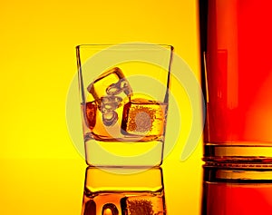 One glass of whiskey with ice cubes near bottle on table with reflection, warm yellow tint atmosphere