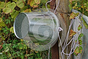 One gray metal zinc bucket hangs on a wooden pole with a white rope near a well