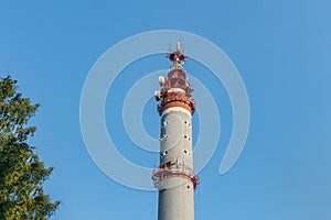 One gray and red Radio relay tower with group of different antennas and green birch tree on the blue sky background photo