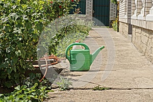 One green plastic watering can and a red watering hose on a gray concrete pavement