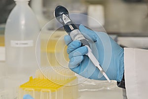 One hand with blue nitril gloves holding a micropipette in a laboratory with other equipment in the background.
