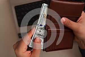 One hundred dollar banknotes in bundle protruded from leather brown wallet in men`s hand on laptop background