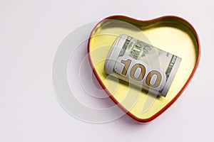 One hundred dollars in a gold heart shaped box with a red outline. Concept for the holiday St. Valentine`s Day, a gift