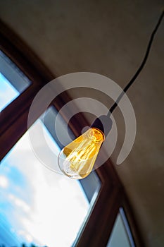 One incandescent bulb near semicircular wooden window with blue cloudy sky overlooking. Bright glowing filament close up