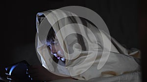 One kids using tablet pc under blanket at night. The boy plays computer games