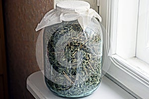 One large glass jar full of dry green tea stands on a white table by the window