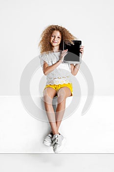 One little cute curly girl in casual clothes sitting on big box with digital tablet, gadget isolated on white studio
