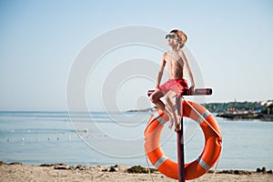 Summer concept of one little kid in red shorts and sunglasses sitting on beach with lifebuoy during tan sunbathe leisure activity