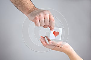 One male hand gives to one female hand a white box with a heart,  simbol of love on a white background, close up