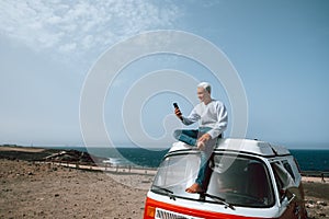 One man alone and isolated sitting on a minivan or camper using his cellphone smiling and looking at it - digital nomad people