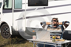 One man digital nomad lifestyle working on alternative outside workplace desk with a camper van motorhome house in background.