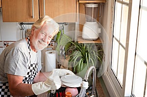 One people senior man white hair and beard wearing apron and gloves washing dishes. Corner of kitchen. Grandfather at work. Bright
