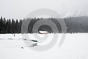 One person dressed in red in a winter landscape. Frozen lake, wooden house and a forest under the snow. Banff National Park, Alber