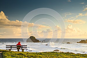 One person sits on bench by ocean, practicing social distancing.