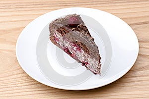 One piece of chocolate cake with vanilla cream and cherry topping served on white plate over light wooden background.