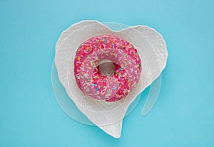 One pink sweet doughnut sits on a white heart-shaped plate on a blue background