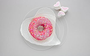 One pink sweet doughnut sits on a white heart shaped plate on a gray background