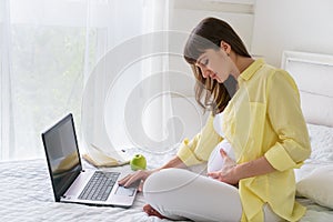 One pregnant european woman working from her home while she is sitting on a white bed at day time