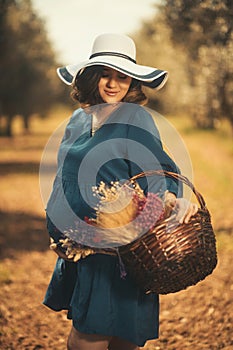 One pregnant woman with a blue dress white hat and a basketfull dry flowers in an olive field with shallow depth of field