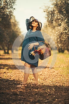 One pregnant woman with a blue dress white hat and a basketfull dry flowers in an olive field with shallow depth of field