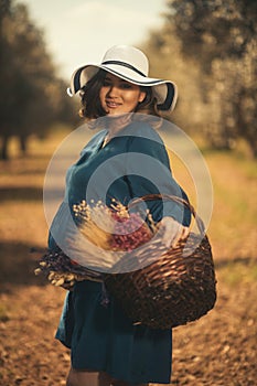 One pregnant woman with a blue dress white hat and a basketfull dry flowers in an olive field with shallow depth of field