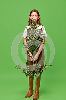One pretty girl with long hair wearing boots holding palm leaf over green background. Child of nature