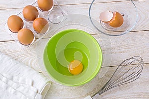 One raw egg in a green bowl, five brown chicken eggs in a transparent plastic tray and  stainless steel whisk on a white wooden
