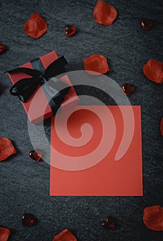 One red box, glass hearts, rose petals and an empty sheet on a black stone background.