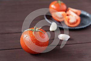 One red ripe tomato and slices of tomato and garlic in the background on a wooden table