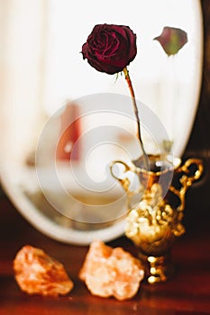 One red wilting rose in a golden vase against an oval mirror and large stones for decor