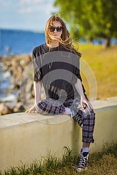 One Relaxing Caucasian Blond Female Girl in Street Clothing Posing Outdoors While Sitted On Stony Fence Outdoors At Sunny Day Near