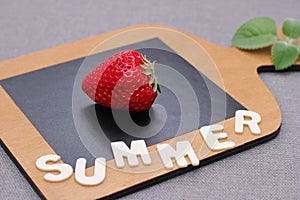 One ripe red fresh strawberry on a wooden chopping board next to the word SUMMER in wooden letters