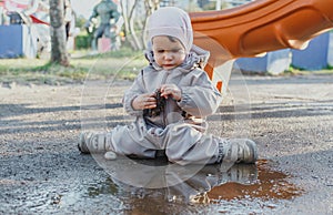 one small child in dirty clothes sits in a puddle on the playground and plays with a cone, the concept of childhood